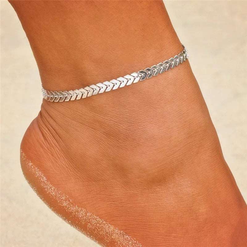 Close-up of an elegant silver herringbone chain Anklet in Gold, worn on a beach with a sandy background.