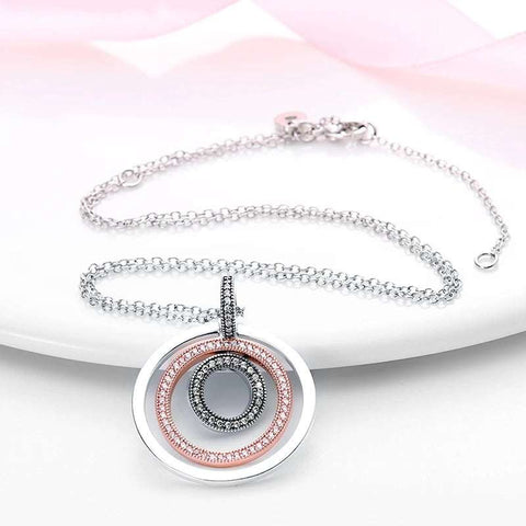 Elegant circular pendant necklace with three concentric rings in silver, rose gold, and diamond-encrusted silver, displayed flat on a white surface with a pink ribbon.