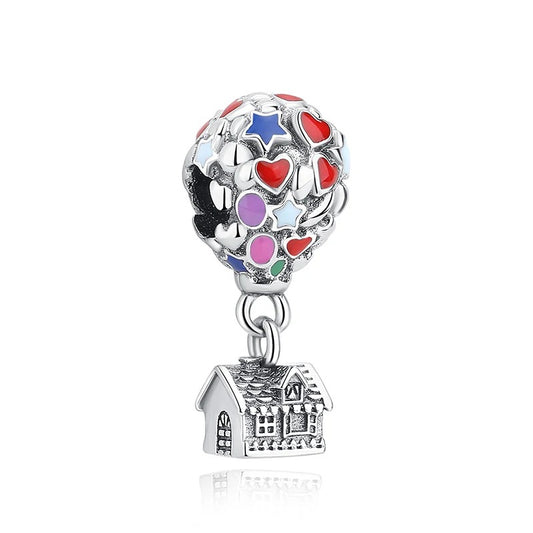 A silver-tone dangle charm featuring a colorful enamel hot air balloon with red hearts, blue and pink stars, and purple circles, suspended above a detailed miniature house charm