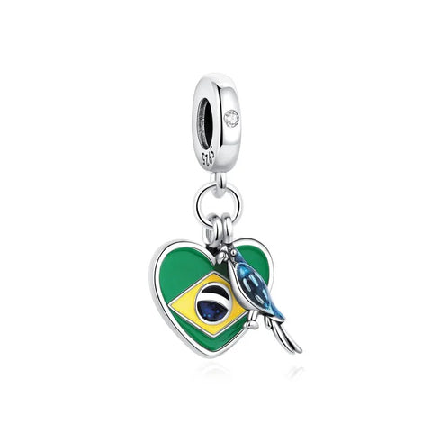 A sterling silver dangle charm featuring a green and yellow enamel heart shaped like the Brazilian flag with a blue enamel macaw parrot perched on it, hanging from a silver clip bail with a clear crystal accent.