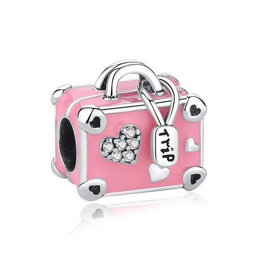 A silver-tone charm shaped like a pink enamel suitcase with heart decorations, a crystal-encrusted heart on the front, and a small luggage tag engraved with the word Trip