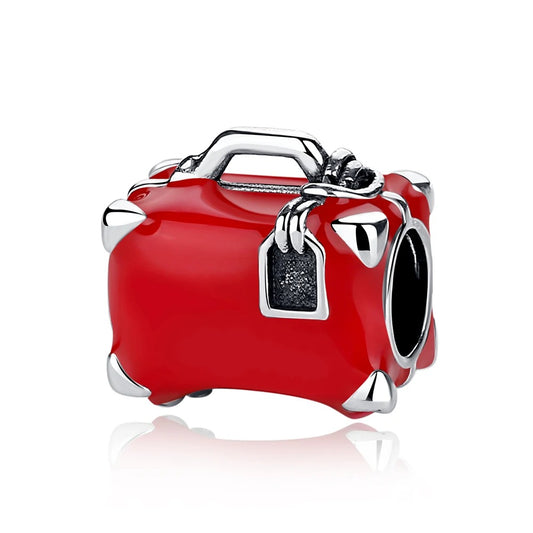 A silver-tone and red enamel miniature suitcase charm with corner guards, carry handle, and luggage tag detail, shown as a bracelet charm