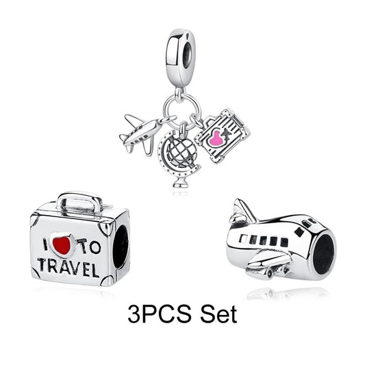 A silver-tone 3-piece travel charm set showing a suitcase bead reading I Love to Travel, an airplane bead, and a dangle charm with miniature plane, globe, and boarding pass with red and pink enamel
